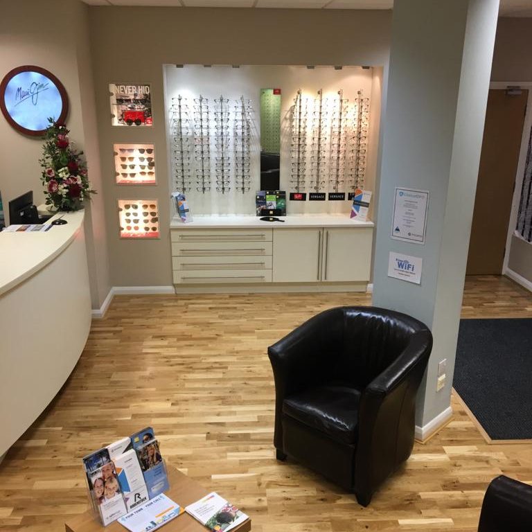 View of the inside of an optician's showing a black chair, reception desk and display stands of glasses.