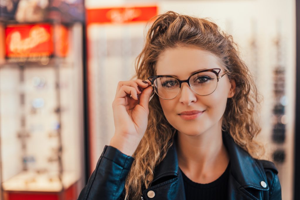 Young woman in a leather jacket holding on to the side of the cat-eye glasses frames she is wearing.