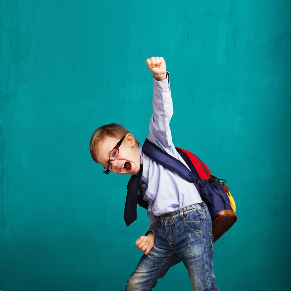 A young boy in jeans, a shirt and tie, wearing a backpack and punching the air.